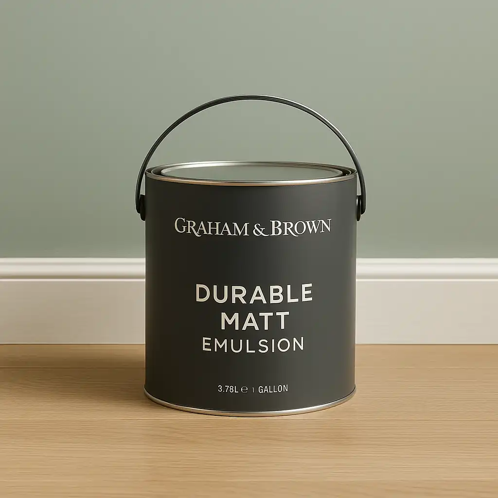 A 1-gallon can of Graham & Brown Durable Matt Emulsion in matte charcoal gray with minimalist white text, placed on a light wood floor against a sage green wall.